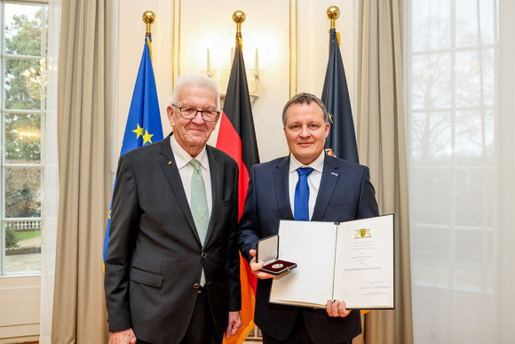Auszeichnung Staufermedaille in Gold für Thomas Speidel Ministerpräsident Winfried Kretschmann (links) und Thomas Speidel (rechts) mit der Staufermedaille in Gold und der Urkunde. Im Hintergrund stehen Fahnen.