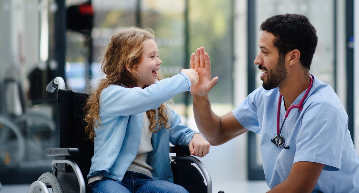 Junger Arzt hat Spaß mit einem kleinen Mädchen im Rollstuhl.
