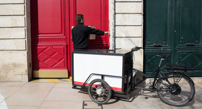 Ein Paketzusteller liefert ein Paket mit einem elektrischen Lastenrad.']