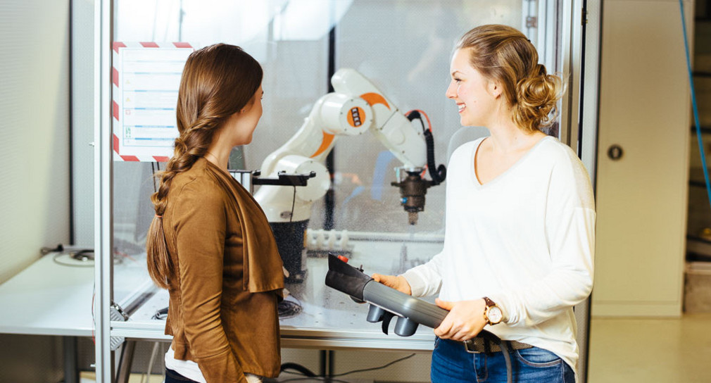 Zwei Frauen stehen vor einem Schaukasten, in dem ein Roboterarm zu sehen ist. 