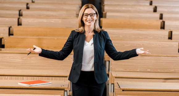 happy female university professor with outstretched hands looking at camera in classroom